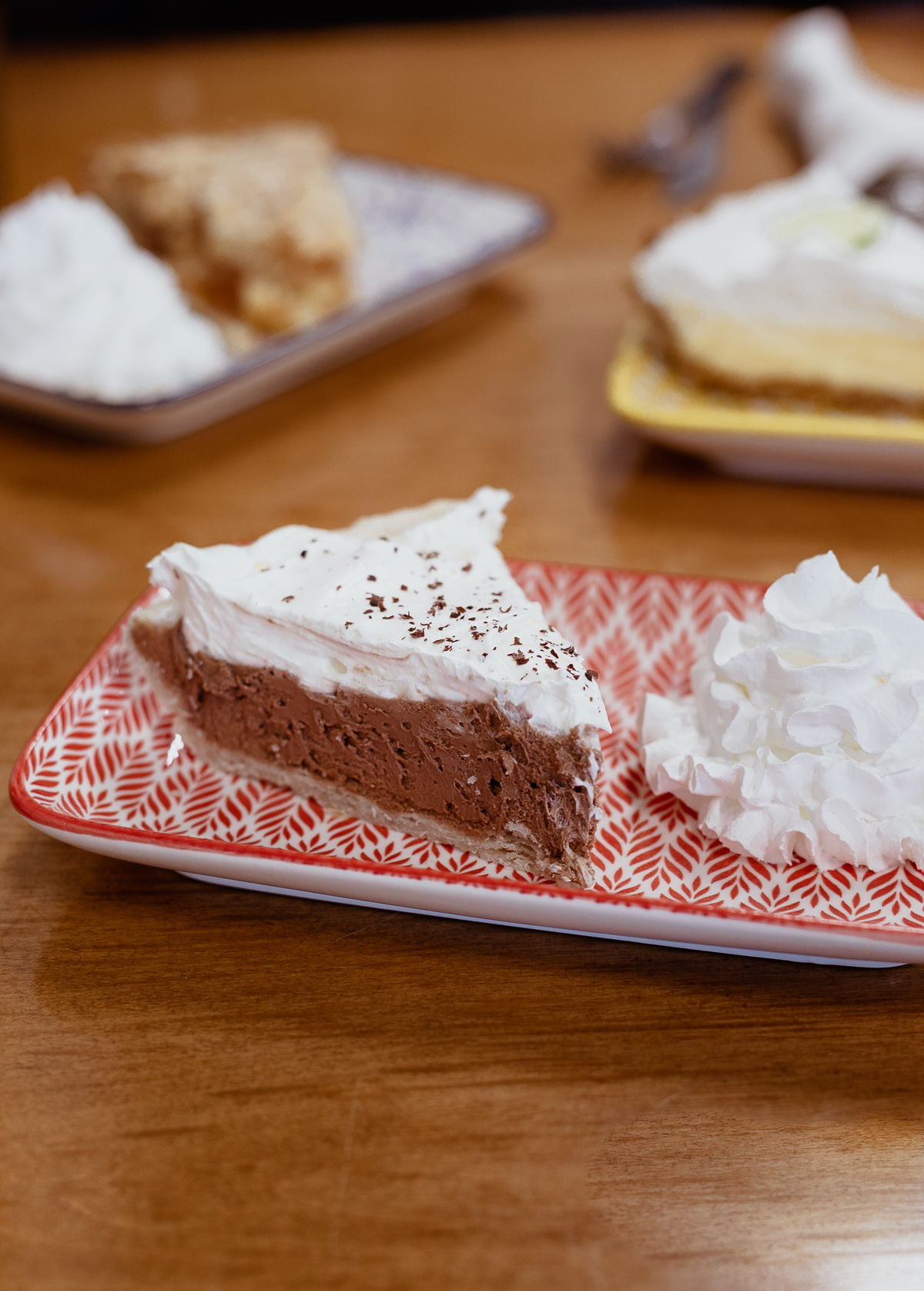 slice of chocolate pie and whipped cream on a red plate with two other slices of pie in the background.